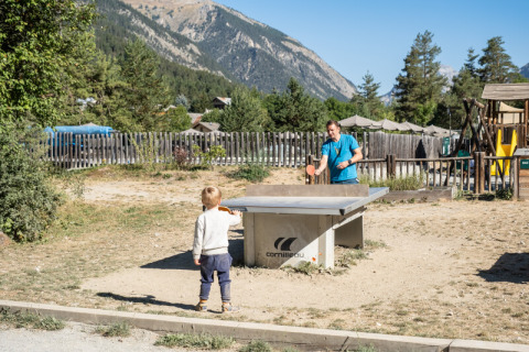 An adult and a child play outdoor table tennis at Huttopia la Clarée holiday park with mountains behind.