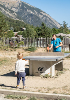 Un adulto y un niño juegan al tenis de mesa al aire libre en Huttopia la Clarée con montañas al fondo.