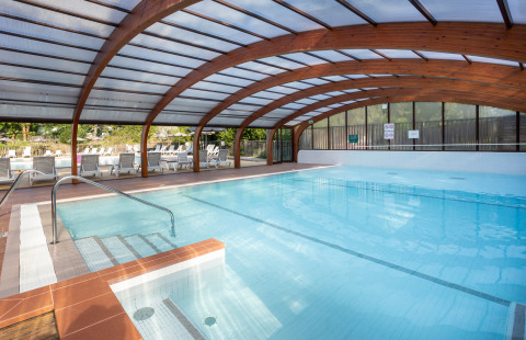 Piscina cubierta con vigas de madera arqueadas y tumbonas exteriores en Camping La Pinède, Excenevex, Francia.