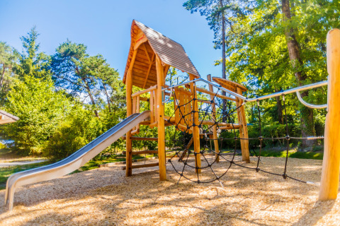 Parque infantil con tobogán y red de escalada rodeado de árboles en Camping La Pinède, Francia.