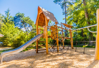 Parque infantil con tobogán y red de escalada rodeado de árboles en Camping La Pinède, Francia.