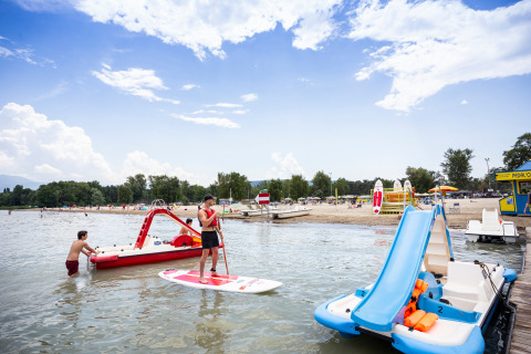 Escena en la playa de Camping La Pinède - Excenevex con hidropedales, paddle surf y bañistas.