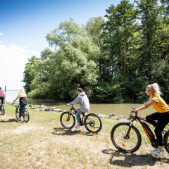 Cuatro personas montan en bicicleta junto a un río cerca de Excenevex-Plage, Francia, en un día soleado.