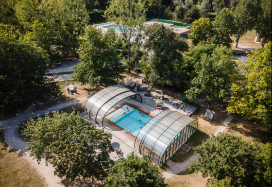 Luftfoto af en overdækket pool og grønne omgivelser i ferieparken Huttopia Les Châteaux i Frankrig.