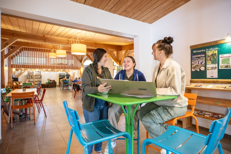 Drie jonge vrouwen werken samen rond een laptop in de lounge van Huttopia Les Châteaux, Frankrijk.