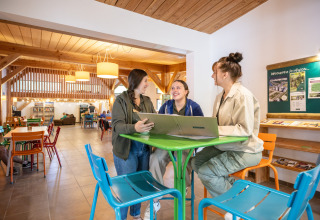 Three young women sit together with a laptop in the communal area of Huttopia Les Châteaux, France.