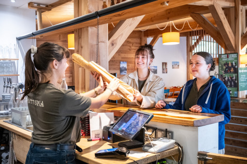 Dos mujeres reciben baguettes frescas de una empleada en el mostrador de Huttopia Les Châteaux, Francia.