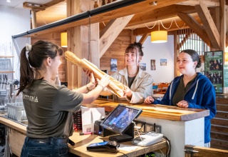 To kvinder modtager baguetter fra en medarbejder bag disken på Huttopia Les Châteaux i Frankrig.