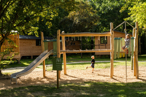 Niños jugando en un parque infantil delante de cabañas de madera en Huttopia Les Châteaux, Centre-Val de Loire.