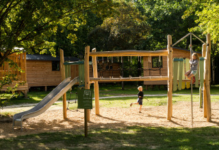 Niños jugando en un parque infantil delante de cabañas de madera en Huttopia Les Châteaux, Centre-Val de Loire.