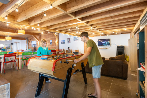 Zwei Kinder spielen Tischfußball in einem hellen Freizeitraum im Huttopia Les Châteaux, Frankreich.