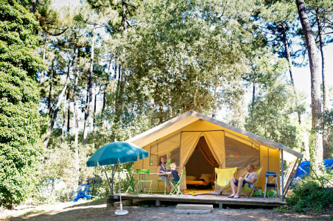 Familia relajándose frente a una tienda safari Bungalow Classic en Huttopia Oléron Les Chênes Verts, rodeada de bosque.