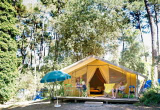 Familia relajándose frente a una tienda safari Bungalow Classic en Huttopia Oléron Les Chênes Verts, rodeada de bosque.