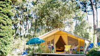 Famiglia che si rilassa davanti a una tenda safari Bungalow Classic a Huttopia Oléron Les Chênes Verts, immersa nel bosco.