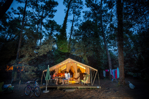 Photo d'une tente safari Bungalow tent Classic éclairée, entourée d'arbres, des gens dînent dehors.