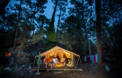 Imagen del Bungalow tent Classic, una tienda safari iluminada en el bosque, personas cenando afuera.