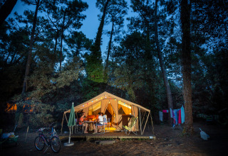 Imagen del Bungalow tent Classic, una tienda safari iluminada en el bosque, personas cenando afuera.