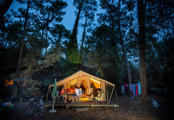 Imagen del Bungalow tent Classic, una tienda safari iluminada en el bosque, personas cenando afuera.