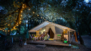 Four people relax in the evening outside a lit safari tent in the woods, surrounded by string lights and bikes.