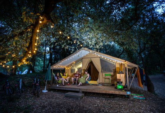Quatre personnes se détendent devant une tente safari illuminée le soir, au cœur de la forêt et de guirlandes.