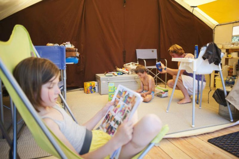 Famille se détend dans une grande tente safari, enfants lisent et jouent à Huttopia Oléron Les Chênes Verts, France.