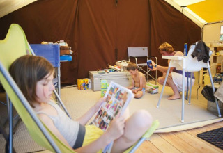Famiglia che si rilassa in una spaziosa tenda safari con bambini che leggono e giocano a Huttopia Oléron Les Chênes Verts, Francia.