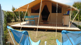 Safari Bungalow Classic-tent met houten terras, blauwe tuinstoelen en twee relaxstoelen op het gras.