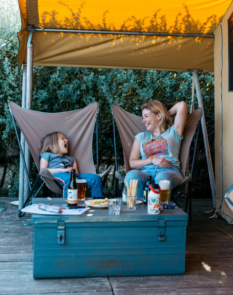 Mamma e figlia ridono e si rilassano in una tenda safari al Huttopia Côte Sauvage, Francia, sotto il telo.