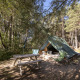 Teepee-teltet Bonaventure midt i en frodig skov, med picnicbord og campingstole i dagslys.