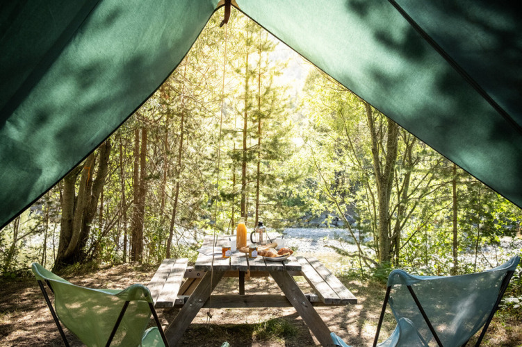 Vista desde una tienda tipi hacia una mesa de picnic con desayuno en un bosque soleado en Huttopia la Clarée, Francia.