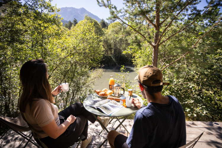 Twee mensen ontbijten aan een buitentafel met uitzicht op een rivier en bergen bij Huttopia la Clarée in Frankrijk.