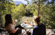 Two people enjoy breakfast at an outdoor table surrounded by trees with a river and mountains at Huttopia la Clarée.