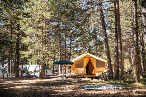 Tienda safari Bungalow tent Classic en bosque, rodeada de árboles, bicicletas, mesas y otras tiendas de campaña.