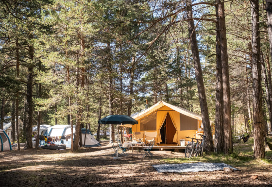 Tienda safari Bungalow tent Classic en bosque, rodeada de árboles, bicicletas, mesas y otras tiendas de campaña.