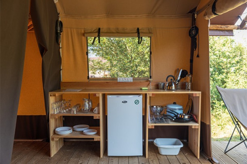 Compacte keuken in een safaritent met houten rekken, servies, een koelkast en kookgerei voor de camping.