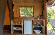 Pequeña cocina en una tienda safari con estantes de madera, platos, nevera y utensilios de camping.