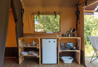 Compacte keuken in een safaritent met houten rekken, servies, een koelkast en kookgerei voor de camping.