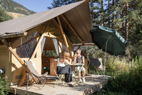Two people relaxing with drinks in front of a Trappeur safari tent, surrounded by greenery and a sun umbrella.