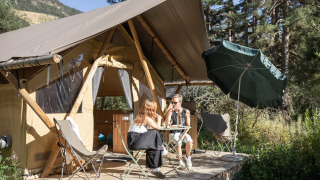 Dos personas disfrutan bebidas fuera de una tienda safari Trappeur, rodeados de naturaleza con sombrilla.