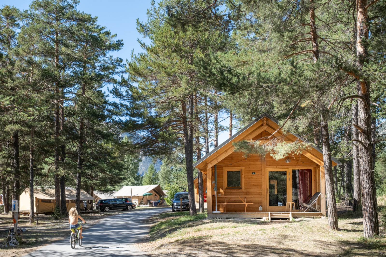 Chalet Montana Vista houten huisje bij Huttopia la Clarée, Frankrijk, tussen bomen en met een fietsend kind.