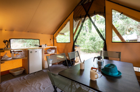 Vista interna di una tenda safari con tavolo da pranzo, sedie e angolo cottura al Camping La Pinède, Francia.