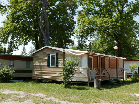 Cabaña tipo mobile home con terraza sombreada y sillas al aire libre, rodeada de árboles y césped.