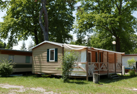 Cabaña tipo mobile home con terraza sombreada y sillas al aire libre, rodeada de árboles y césped.