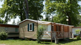 Mobile home cabin with shaded porch and outdoor chairs, surrounded by greenery and tall trees.