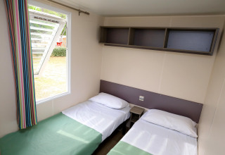 Zwei Einzelbetten mit weißen Bettlaken und offenem Fenster im Mobile Home auf Camping La Pinède, Excenevex, Frankreich.