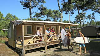 Grupo de jóvenes relajándose en una cabaña móvil, rodeados de árboles y dos con tablas de surf.