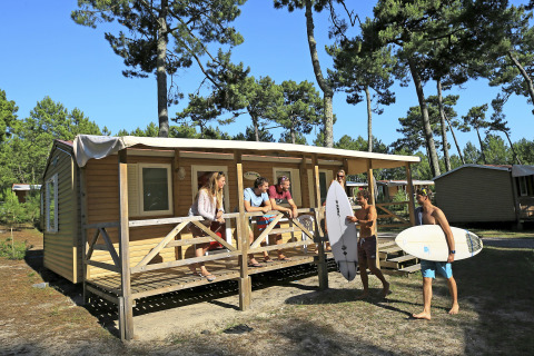 Groupe de jeunes devant un mobil-home en bois, dans une forêt, deux portent des planches de surf.
