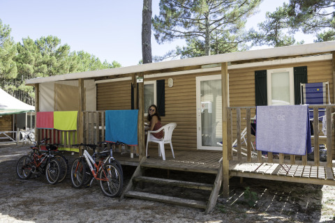 Mujer sentada en el porche de una casa móvil en Camping La Pinède, Excenevex, Francia, con bicicletas y toallas.