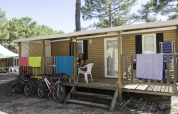 Mujer sentada en el porche de una casa móvil en Camping La Pinède, Excenevex, Francia, con bicicletas y toallas.