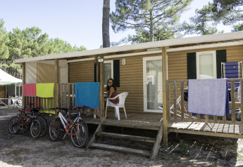 Mujer sentada en el porche de una casa móvil en Camping La Pinède, Excenevex, Francia, con bicicletas y toallas.